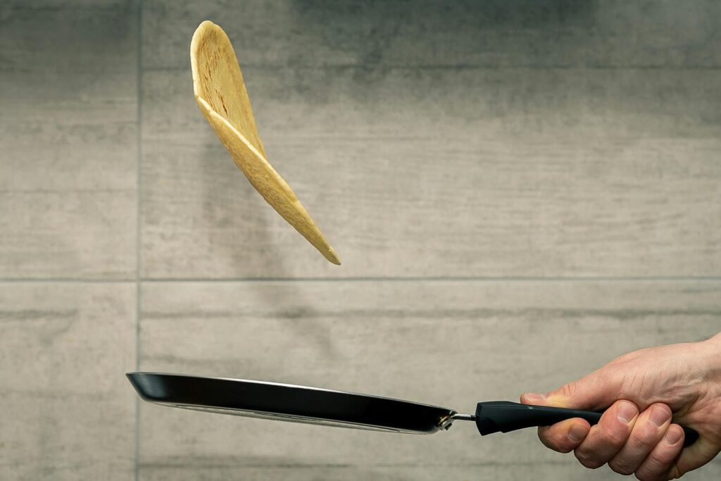 A hand skillfully flips a pancake mid-air in a frying pan, showcasing cooking expertise.