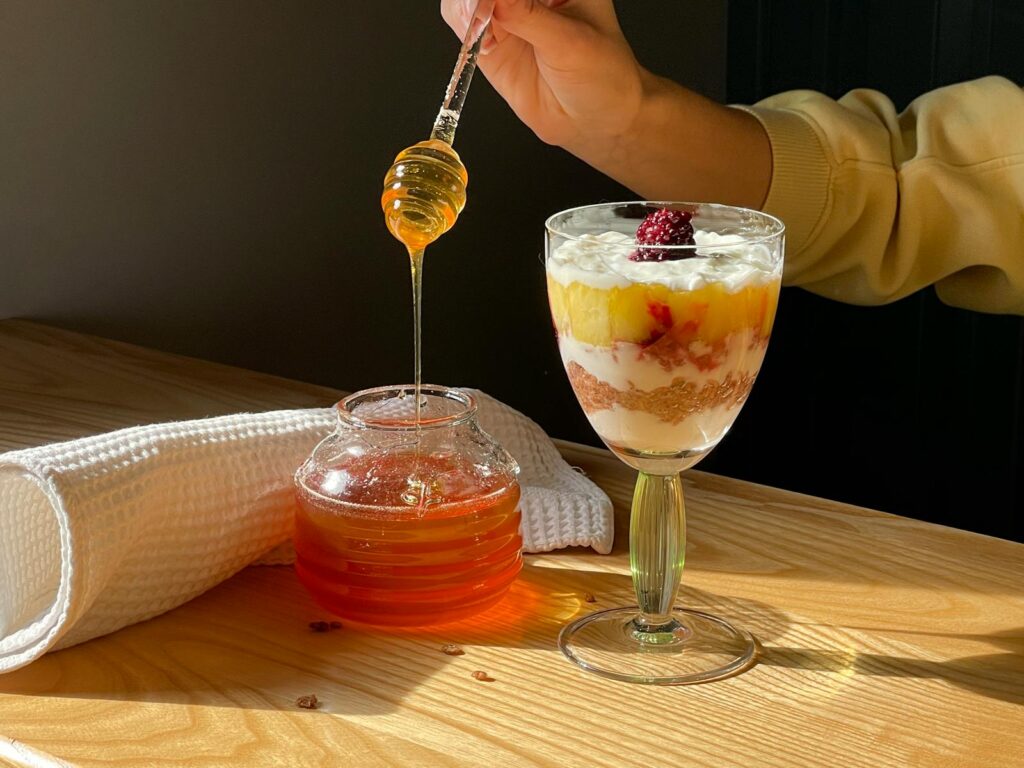 A hand drizzles honey over a fruit parfait in warm sunlight, creating a delicious dessert scene.