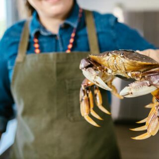 Woman in apron holding a live crab in a kitchen setting, showcasing fresh seafood.
