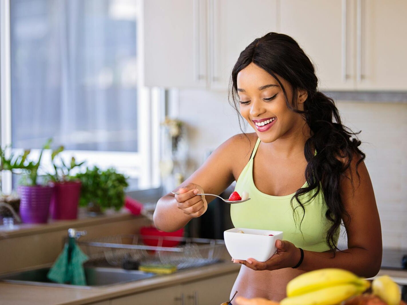 Smiling woman eating a nutritious fruit bowl in a bright kitchen setting.