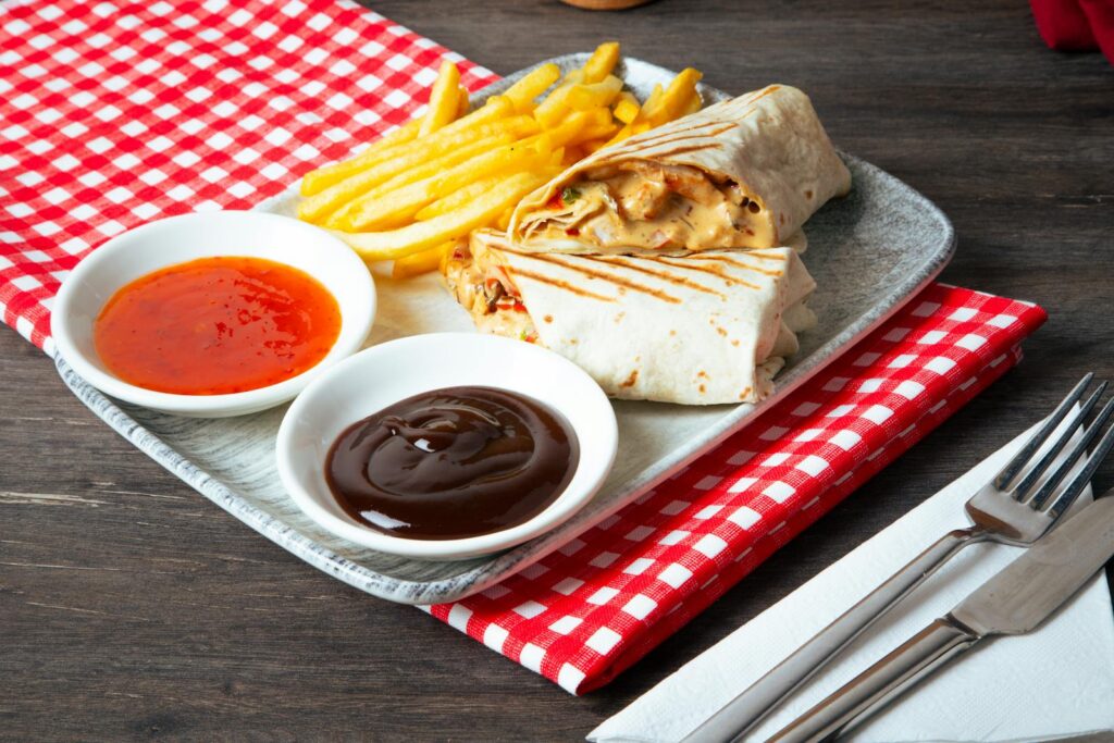 Close-up of a tasty fast food meal including wraps, french fries, and dipping sauces on a checkered cloth.