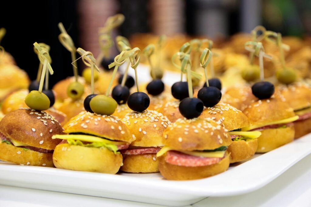 A tray of gourmet mini burgers topped with green and black olives, perfect for catering events.