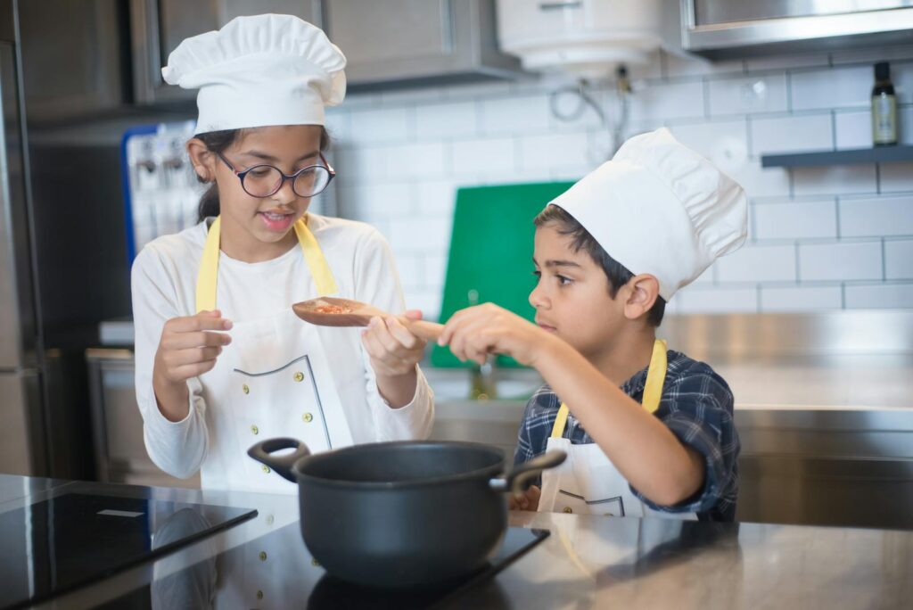 Two young children in chef hats cooking together in a modern kitchen setting.