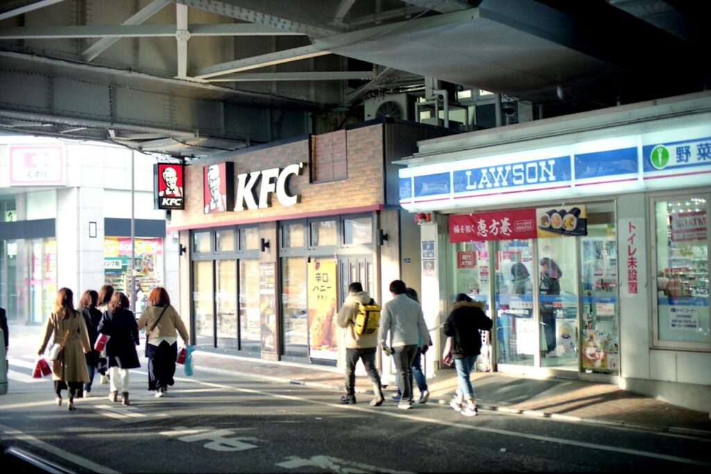 People walking past KFC and Lawson stores under an overpass in Tokyo.