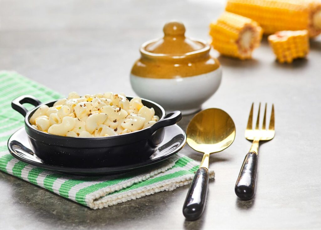 Delicious creamy macaroni with corn served in a black bowl alongside cutlery on a checkered cloth.