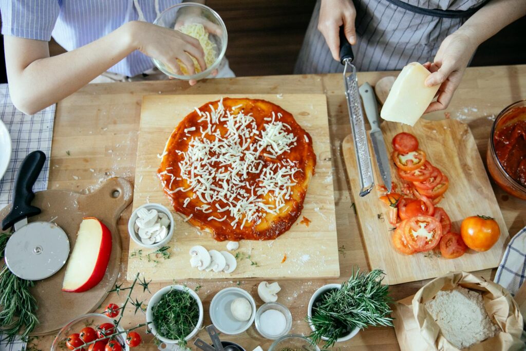 Overhead view of hands preparing a homemade pizza with fresh ingredients like cheese, tomatoes, and herbs.